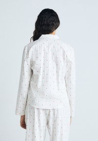 Camicia bianca a maniche lunghe con righe verticali, piccoli cuori rossi sparsi e colletto. Tessuto morbido con una vestibilità rilassata.