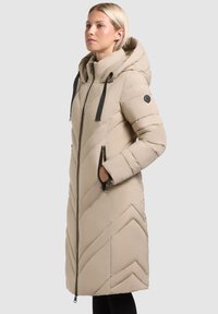 Beige polstret jakke med hætte, med et chevron mønster, sorte lynlåslommer og snoretræk på hætten og den forreste lynlås.
