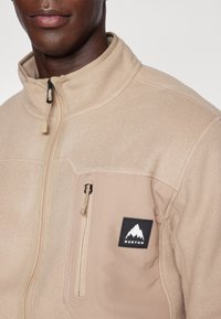 Beige fleecejakke med høy krage, en glidelås-lomme på brystet, og et svart merke med en hvit fjelllogo på venstre bryst.