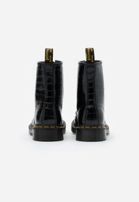 Para czarnych lakierowanych botków za kostkę z wzorem krokodyla, żółtym przeszyciem i czarnymi uchwytami do zakupu z żółtym napisem, widoczna z tyłu.