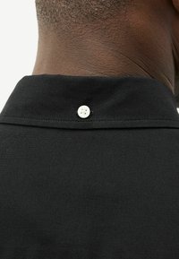 Cuello de camisa negro con una tela texturizada, que presenta un botón blanco en la parte posterior, destacando un diseño simple y líneas limpias.