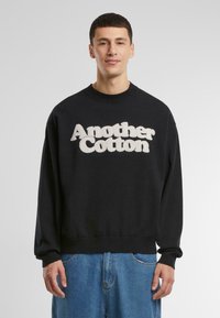 Sudadera de algodón negra con un logo bordado blanco y peludo "Another Cotton" en la parte frontal. Cuello redondo y puños acanalados.