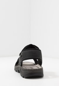 Svart sandal med en texturerad ovandel, justerbara remmar och en vadderad sula. Har kontrasterande vit sömnad och ett robust grepp.