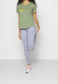 Ljust grönt sport-T-shirt med en gul Nike-logotyp, kombinerat med ljuslila leggings med dragkedjor och vita sneakers med rosa detaljer.