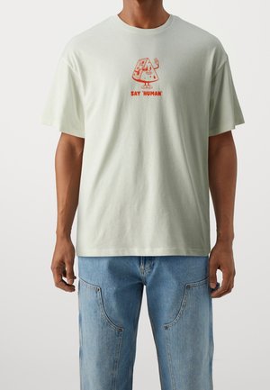 Homme portant un t-shirt vert clair avec un personnage de pizza rouge de dessin animé et le texte « DIS 'HUMAIN' », associé à un jean bleu avec de grandes poches devant.