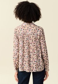 Langärmlige Bluse mit floralem Muster auf hellbeigem Hintergrund, ausgestattet mit verschiedenen bunten Blumenmustern, einem gefältelten Rückendesign und klassischem Kragen.