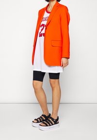 Orange kavaj över en vit mesh baskettröja med nummer 23, svarta cykelshorts och svarta plattformssandaler med remmar.