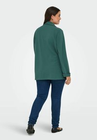 Žalia pūkinė striukė su išpjauta apykakle, ilgomis rankovėmis ir laisvu siluetu, derinama su tamsiai mėlynomis džinsinėmis keln�ėmis ir juodais butais.