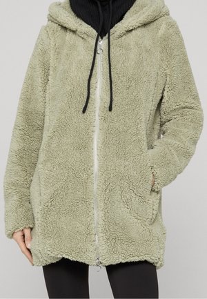 Veste zippée en polaire sherpa vert clair avec capuche et poches latérales, portée sur un sweat à capuche noir et des leggings noirs.