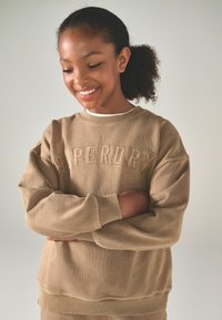 Tanfärgad sweatshirt med ribbade ärmslut och nederkant. Har broderad "SUPERDRY" i en något mörkare nyans. Mjuk textur, avslappnad passform.