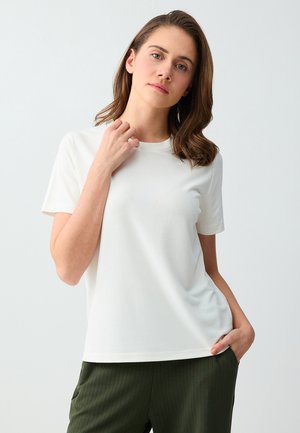 Jeune femme en chemise blanche à manches courtes et pantalon vert foncé, touchant le col d'une main, debout devant un fond clair uni.