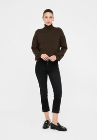 Vrouw in een donkerbruine coltrui, zwarte cropped jeans met opgevouwen zoom, zwarte loafers en gouden oorbellen, staand tegen een witte achtergrond.