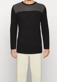Pull en tricot avec un corps noir et un panneau supérieur gris, texture côtelée, col rond et manches longues. Design épuré avec un branding subtil.
