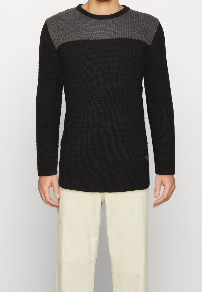 Pull en tricot avec un corps noir et un panneau supérieur gris, texture côtelée, col rond et manches longues. Design épuré avec un branding subtil.