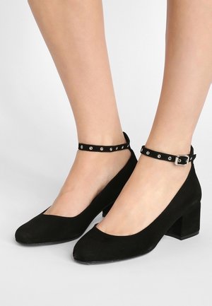 Chaussures à talons hauts en suède noir avec des bouts ronds et un talon bloc. Bride de cheville ornée de petits clous en argent.