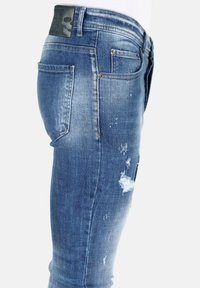 Slim fit blauwe denim jeans met versleten patches en verfspetters; heeft een achterzak en een leren label in het donker op de tailleband.