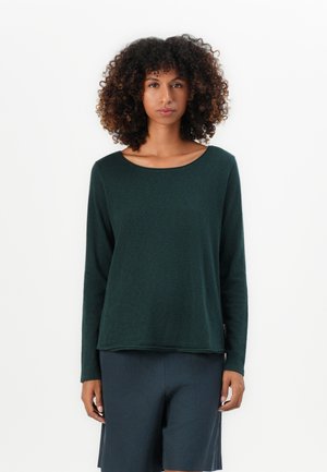 Donna con capelli ricci che indossa una maglia a maniche lunghe verde scuro e pantaloni corti blu scuro, in piedi di fronte a uno sfondo bianco uniforme.