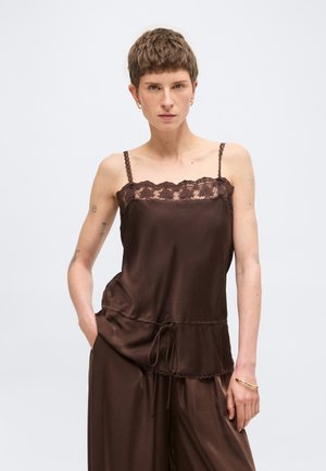 Kvinde med kort hår iført brun satin camisole med blondekant og matchende bukser, poserer med hånd i lommen mod hvid baggrund.