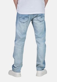 Man i ljusblå denimjeans med sömnad på bakfickorna, vita sneakers och en vit t-shirt, stående mot en neutral bakgrund.