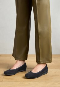 Balerini din suede negru, cu un design cu vârf rotunjit, asortați cu pantaloni verzi măslinii, largi, expuși pe un podea din lemn.
