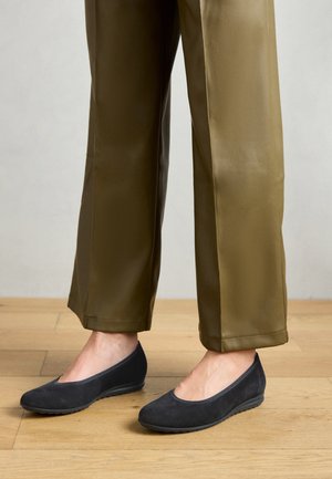 Ballet flats de camurça preta com um design de bico arredondado, combinadas com umas calças largas de cor verde-azeitona, exibidas sobre um chão de madeira.