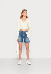 Top cropped amarelo-claro com mangas longas, combinado com calções de ganga azuis. A modelo segura uma pequena bolsa branca e usa ténis brancos.