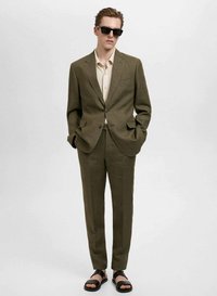 Costum verde măsliniu cu un sacou realizat pe comandă și pantaloni drepti, asortat cu o cămașă bej deschis și ochelari de soare negri. Sandalele negre completează ținuta.