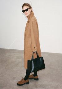 Manteau en laine de chameau avec col montant et boutons sur le devant, sac à main en cuir noir, collants noirs et chaussures en suède marron à lacets avec semelles épaisses.