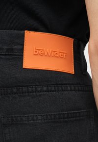Κοντινό πλάνο της μέσης μαύρου τζιν, με ραμμένη πορτοκαλί ορθογώνια ετικέτα που εμφανίζει το όνομα της μάρκας "bewilder" με ανάγλυφα γράμματα.