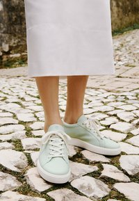 Sneaker in pelle verde con lacci bianchi e suole in gomma striata, posizionati su un pavimento di ciottoli e una gonna bianca leggera.