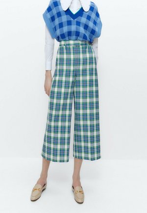 Model draagt een hooggetailleerde, wijd uitlopende, cropped geruite broek in groen, blauw en wit, gecombineerd met een blauw geblokt gebreid vest en beige loafers.