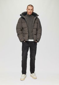 Veste matelassée grise avec capuche, pull en maille texturée en dessous, jeans foncés et baskets claires. Présenté sur un fond neutre.