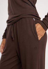 Ruskea ribbikuvioinen oloasu, joka sisältää pitkähihisen paidan ja joustavyötäröiset housut sivutaskuilla sekä brodeeratun logoelementin.