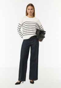 Witte trui met marineblauwe strepen, gecombineerd met donkerblauwe wijde jeans met contrasterende steken. Zwarte handtas en puntige hakken maken de look af.