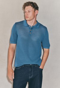 Gestricktes blaues Poloshirt mit kurzen Ärmeln, das einen Kragen und drei silberne Knöpfe hat, kombiniert mit dunklen Jeans aus Denim.