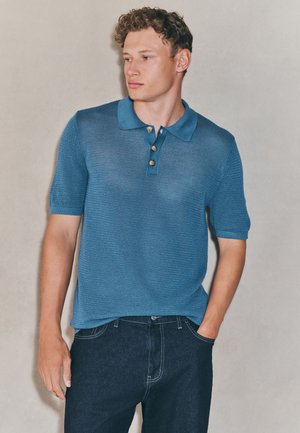 Gebreid blauw poloshirt met korte mouwen, voorzien van een kraag en drie zilveren knopen, gecombineerd met donkere spijkerbroek.
