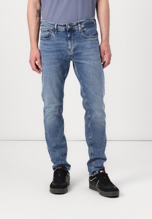 Mann, der eine Slim-Fit hellblaue Jeans und schwarze Sneakers trägt und vor einem weißen Hintergrund steht.
