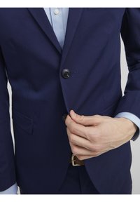Granatowa marynarka z dwoma guzikami, klapami w kształcie notcha i boczną kieszenią. Gładki materiał, dopasowany krój, zestawiona z jasnoniebieską koszulą pod spodem.