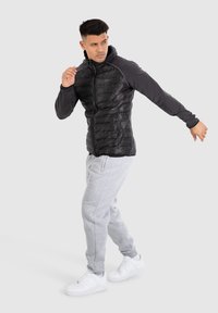 Schwarze Kapuzenjacke mit gestepptem Design, kombiniert mit grauen Jogginghosen. Das Model steht in einer dynamischen Pose und trägt weiße Sneakers.