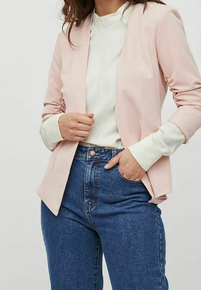 Ljusrosa blazer med skräddarsydd design, använd över en vit topp. Kombinerad med högmidjade blåjeans, som har en slät textur och klassisk passform.