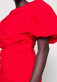 Eine dunkelhäutige Person trägt ein lebhaft rotes Kleid mit kurzen, puffigen Ärmeln und gerafften Stoffdetails an der Seite.