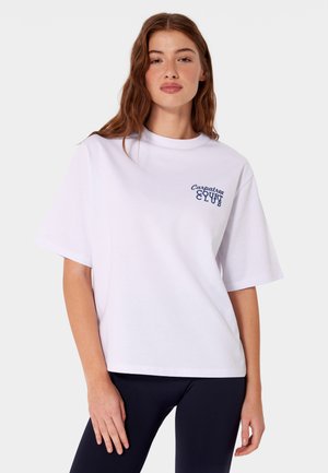 Młoda kobieta z długimi brązowymi włosami, ubrana w białą, oversize koszulkę z napisem „Carpatree Court Club” na lewej piersi i czarne spodnie.