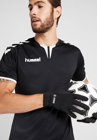 Maillot de football noir avec des accents et un logo blancs, associé à des gants de gardien de but noirs, tenant un ballon de football noir et blanc à motifs.