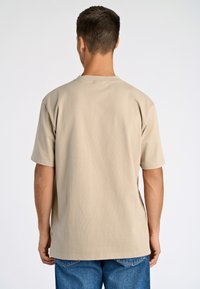 Beige lyhythihainen T-paita, joka on valmistettu puuvillasekoitteesta, jossa on pyöreä kaula-aukko ja rento istuvuus, yhdistettynä sinisiin farkkuihin.