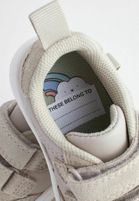 Helle Kindersneaker aus einer Kombination von Leder und Wildleder, ausgestattet mit Klettverschlüssen und einer bunten, gemusterten Innensohle mit einem Etikett.