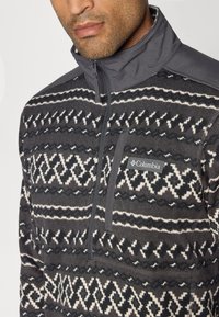 Pull en polaire avec col zippé gris, présentant un motif géométrique noir et blanc, et une poche sur la poitrine avec un détail de logo.