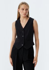 Schwarze taillierte Weste mit tiefem V-Ausschnitt, fünf Knöpfen und zwei Fronttaschen. Glattes Material mit maßgeschneiderter Passform, getragen über schwarzen Hosen.