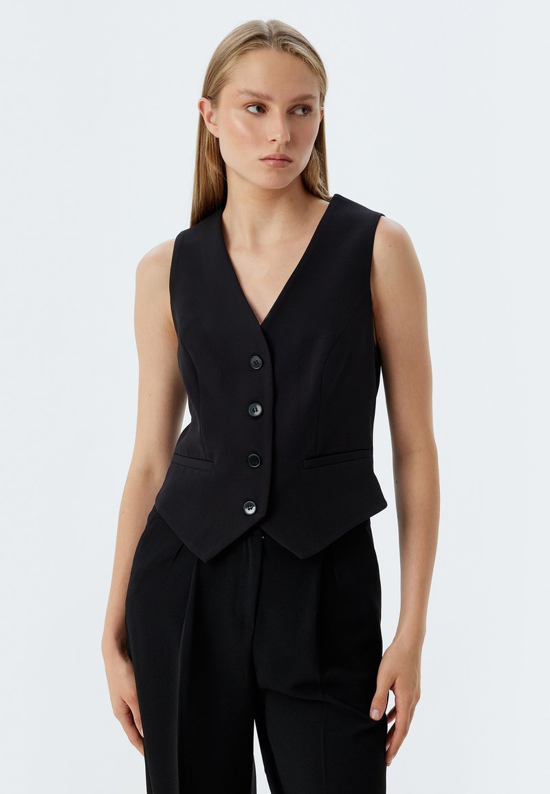 Schwarze taillierte Weste mit tiefem V-Ausschnitt, fünf Knöpfen und zwei Fronttaschen. Glattes Material mit maßgeschneiderter Passform, getragen über schwarzen Hosen.