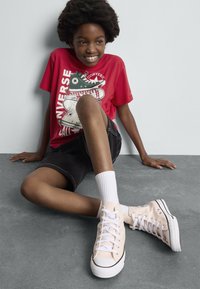 Beige lerret høye sneakers med hvite snører og svarte detaljer, brukt med svarte shorts og en rød grafisk t-skjorte, hvite sokker.
