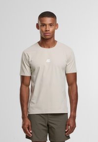 Uomo con capelli corti che indossa una camicia a maniche corte beige chiaro e pantaloni corti verde oliva scuro, in piedi davanti a uno sfondo chiaro e uniforme.